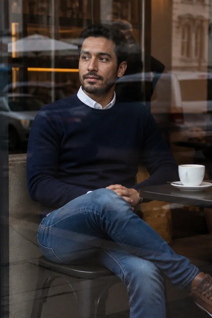 Mid blue jeans with navy sweater over white collared shirt and brown sneakers